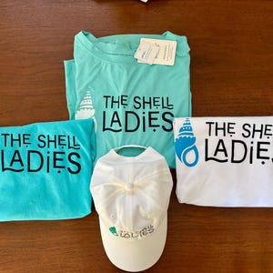 May include: An assortment of apparel featuring the text "THE SHELL LADIES" and a seashell graphic. Items include two turquoise t-shirts, a white t-shirt, and a white baseball cap. The items are displayed on a brown wooden surface.