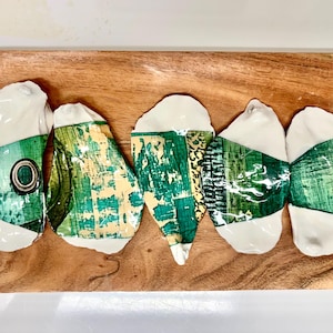 May include: Five ceramic fish-shaped serving dishes with white and green abstract designs, arranged on a wooden cutting board. The fish have a white base with green and yellow patterned accents.