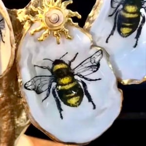 May include: Close-up of decorative bee-themed ornaments. Each ornament features a white, irregular-shaped base with a detailed illustration of a bee in black and yellow. The ornaments are accented with gold-colored embellishments and a sun-shaped detail.