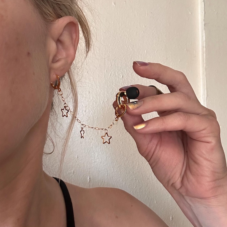 May include: A gold-toned earring featuring a small hoop attached to a delicate chain with three dangling open star charms. The chain connects to a second gold hoop that holds a single matte black spherical bead, creating a layered earring design.