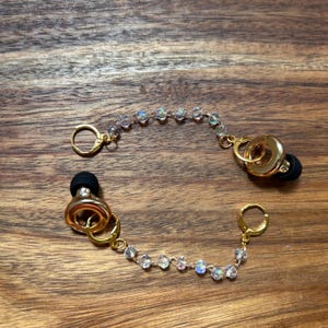 May include: A pair of gold-toned earbud accessories featuring a chain of iridescent, faceted beads. Each piece includes a gold-toned circular ring, a small gold-toned hoop clasp, and a black silicone earbud tip attached to the central metal ring.
