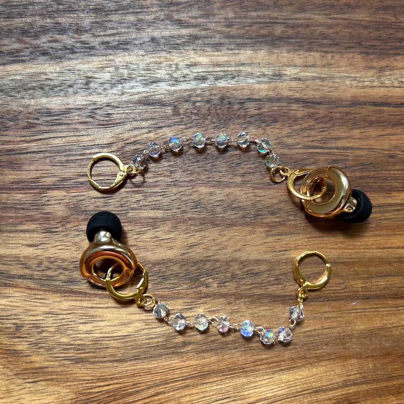 May include: A pair of gold-toned earbud accessories featuring a chain of iridescent, faceted beads. Each piece includes a gold-toned circular ring, a small gold-toned hoop clasp, and a black silicone earbud tip attached to the central metal ring.