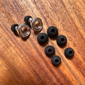 May include: A pair of silver-toned circular earplugs with black silicone tips, accompanied by five additional black silicone replacement tips of varying sizes, arranged in a cluster on a wooden surface with a visible grain pattern.