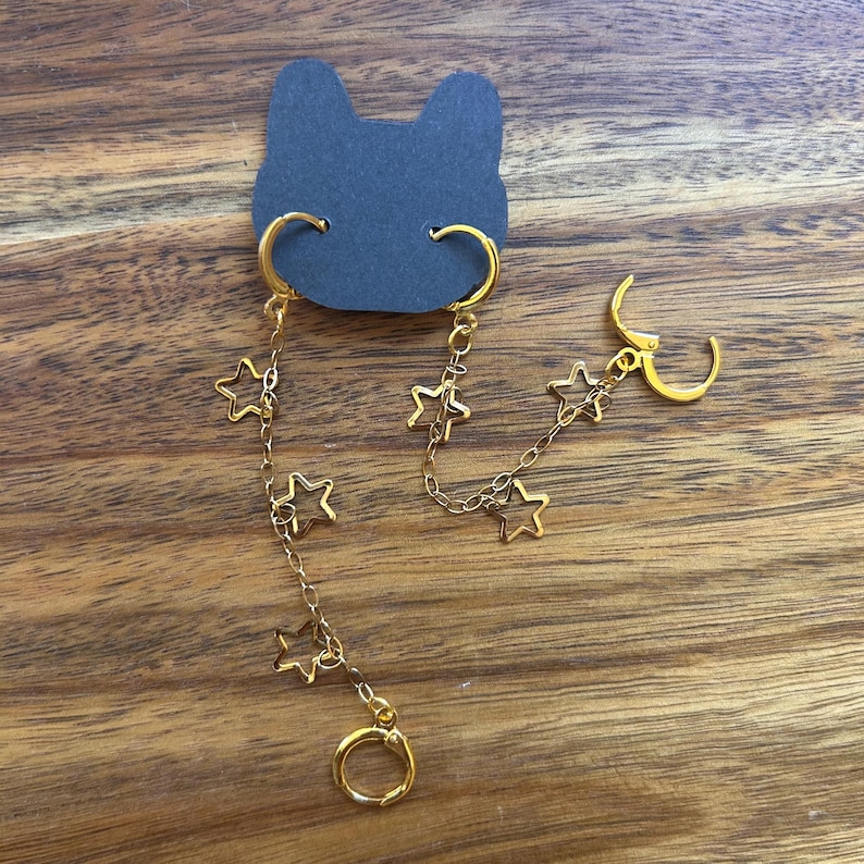 May include: A pair of gold-toned earrings connected by a delicate chain featuring multiple open star-shaped charms. Each earring consists of a circular hoop clasp attached to a chain segment, with the stars spaced evenly along the gold-colored metal links.