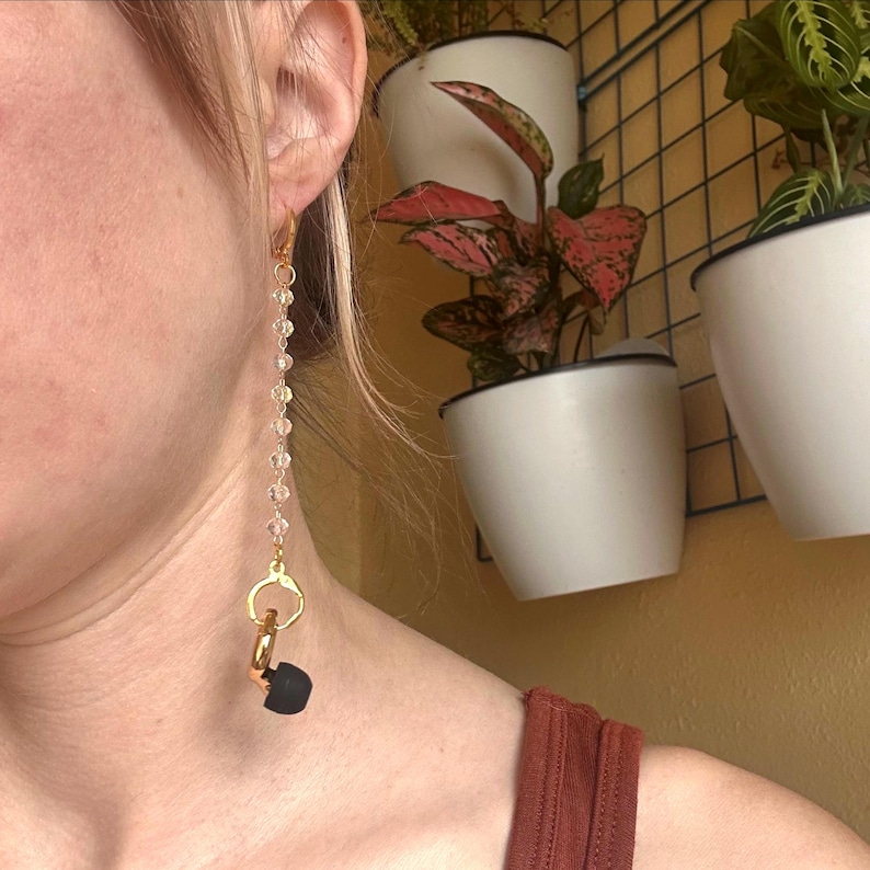 May include: A long dangle earring featuring a gold-toned metal chain adorned with small, clear, faceted crystal beads. The chain connects to a gold-toned metal ring that holds a single, small, matte black earplug at the bottom of the design.