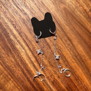 May include: A pair of silver-toned hoop earrings connected by delicate chains. Each earring features a small, four-pointed star charm set with a clear stone, along with additional small clear stone accents. The earrings are attached to a black cat-shaped card.