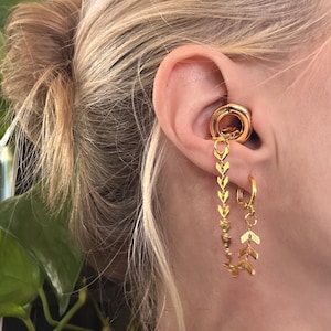 May include: A gold-toned ear cuff featuring a smooth circular ring that sits in the conch, connected by a delicate chain with chevron-shaped links to a small hoop earring in the lobe, creating a layered jewelry piece for the ear.
