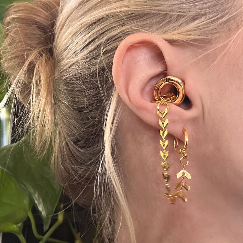 May include: A gold-toned ear cuff featuring a smooth circular ring that sits in the conch, connected by a delicate chain with chevron-shaped links to a small hoop earring in the lobe, creating a layered jewelry piece for the ear.