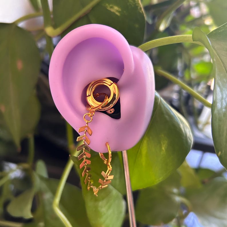 May include: A gold-toned ear cuff and hoop earring set connected by a delicate chain featuring a repeating chevron pattern. The jewelry is displayed on a light purple silicone ear model, showcasing the linked design and polished metallic finish.