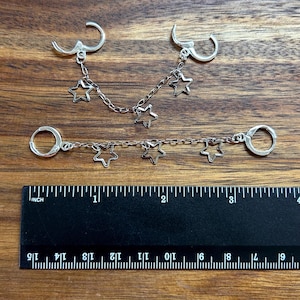 May include: Two silver-toned chain earrings featuring open star charms. The top earring has a curved chain with three stars, while the bottom earring has a straight chain with three stars. A black ruler shows the items are approximately 3.5 inches long.