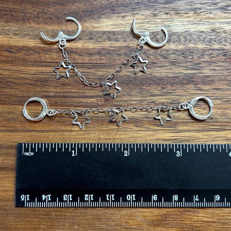 May include: Two silver-toned chain earrings featuring open star charms. The top earring has a curved chain with three stars, while the bottom earring has a straight chain with three stars. A black ruler shows the items are approximately 3.5 inches long.