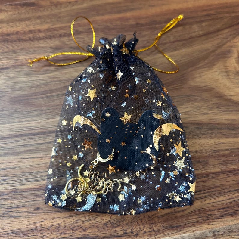 May include: A black sheer drawstring pouch featuring a metallic gold and silver celestial pattern of stars and crescent moons. A black fabric bat-shaped cutout is attached to the front, adorned with a dangling gold-tone metal charm chain.