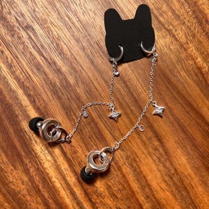 May include: A silver-toned earbud chain accessory featuring two circular connectors with black silicone tips, attached to a black cat-shaped display card. The delicate chain is adorned with small clear crystals and two four-pointed star charms.