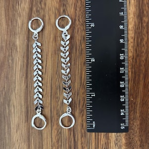 May include: A pair of silver-toned earrings featuring a vertical chain of chevron-shaped links connected by two small circular hoops. A black ruler is positioned next to the earrings, showing a total length of approximately 3.5 inches.