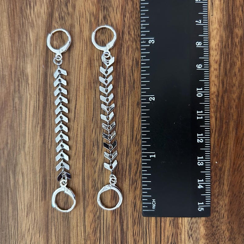 May include: A pair of silver-toned earrings featuring a vertical chain of chevron-shaped links connected by two small circular hoops. A black ruler is positioned next to the earrings, showing a total length of approximately 3.5 inches.