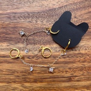 May include: A pair of gold-toned hoop earrings connected by delicate chains. Each earring features a small star-shaped charm with a clear crystal center and additional small crystal dangles. The jewelry is attached to a black cat-shaped display card.