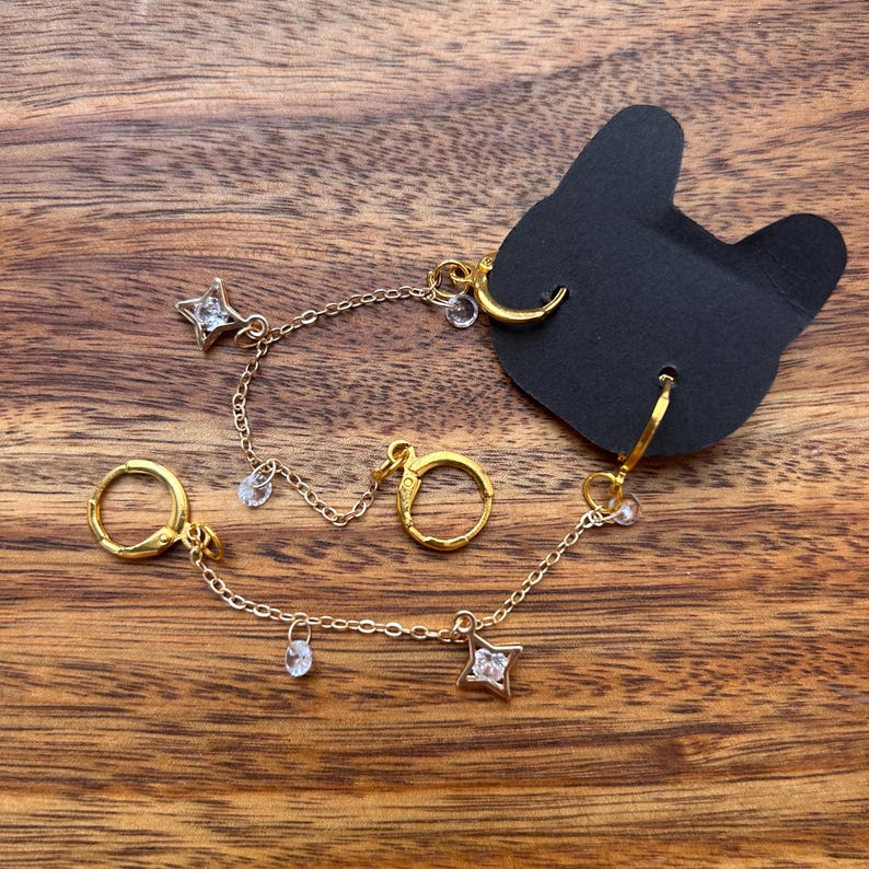 May include: A pair of gold-toned hoop earrings connected by delicate chains. Each earring features a small star-shaped charm with a clear crystal center and additional small crystal dangles. The jewelry is attached to a black cat-shaped display card.