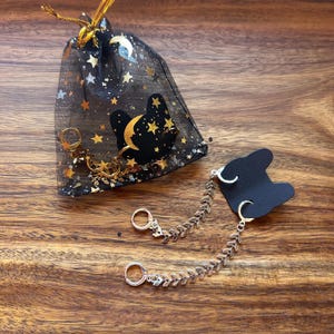 May include: A black sheer drawstring pouch decorated with gold and silver stars and moons, accompanied by two silver-toned earrings. Each earring features a small hoop connected to a long, articulated chain with a chevron-patterned design.