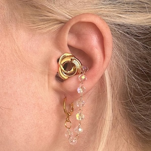 May include: A gold-toned ear cuff featuring a circular knot design positioned in the conch, connected by a delicate chain of small, clear, faceted crystal beads to a gold-toned hoop earring worn in the lower earlobe.
