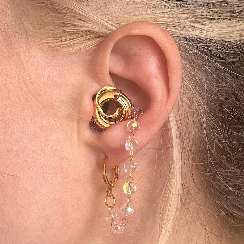 May include: A gold-toned ear cuff featuring a circular knot design positioned in the conch, connected by a delicate chain of small, clear, faceted crystal beads to a gold-toned hoop earring worn in the lower earlobe.