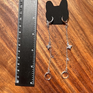 May include: A pair of silver-toned dangle earrings featuring a chain design with a small star-shaped charm and a clear stone, attached to a black cat-shaped display card. A black ruler shows the earrings measure approximately 5.5 inches in length.