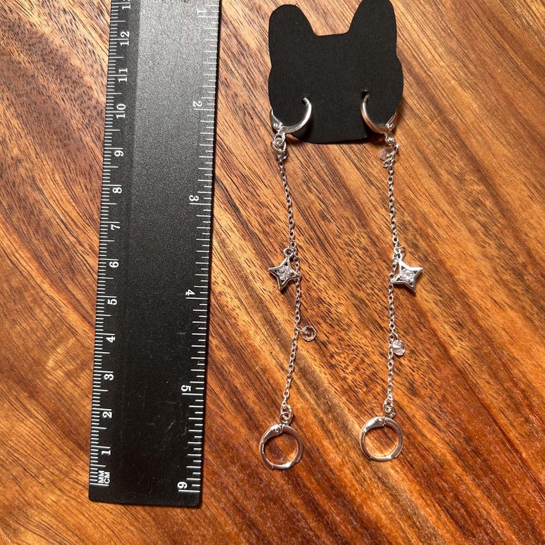 May include: A pair of silver-toned dangle earrings featuring a chain design with a small star-shaped charm and a clear stone, attached to a black cat-shaped display card. A black ruler shows the earrings measure approximately 5.5 inches in length.