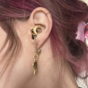 Op de afbeelding: Een goudkleurige oorsieradenset bestaande uit een cirkelvormige oorring in het conch-gebied, verbonden door een fijne metalen ketting met een kleinere oorring in de oorlel, voorzien van een kleine, getextureerde goudkleurige vleugelbedel.
