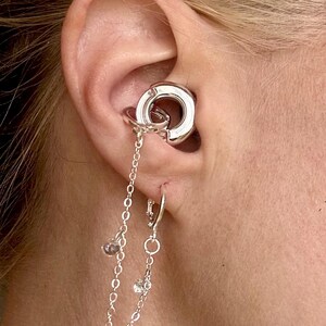 May include: A silver-toned ear cuff and hoop earring set connected by two delicate silver chains. The ear cuff features a thick circular design, while the lower hoop earring includes small dangling charms and clear crystal accents.