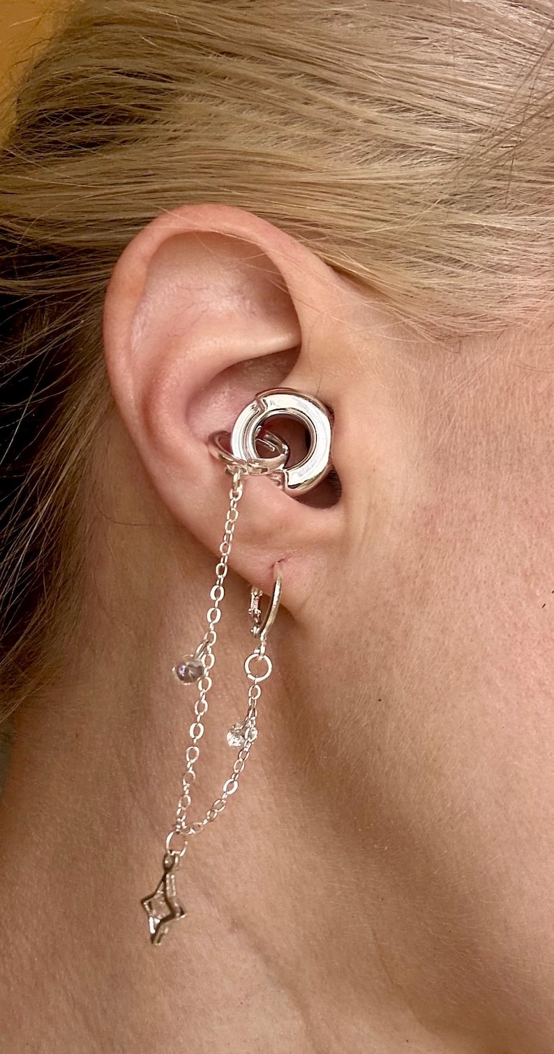 May include: A silver-toned ear cuff and hoop earring set connected by two delicate silver chains. The ear cuff features a thick circular design, while the lower hoop earring includes small dangling charms and clear crystal accents.