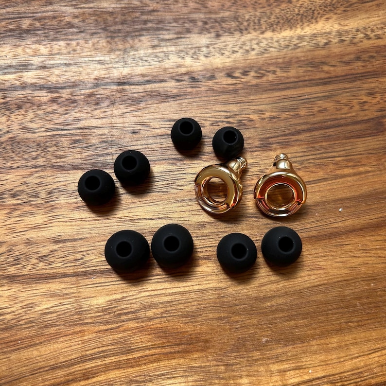 May include: A collection of small components including two polished gold-tone metal circular pieces with threaded stems and eight matte black silicone earbud tips. The gold pieces feature a hollow center and a smooth, reflective finish.