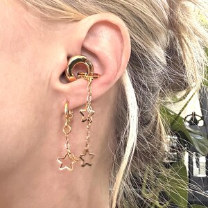 May include: A gold-toned ear cuff featuring a smooth circular band connected to a dangling chain with two open star charms, paired with a matching gold-toned hoop earring that has a single dangling open star charm attached to its base.
