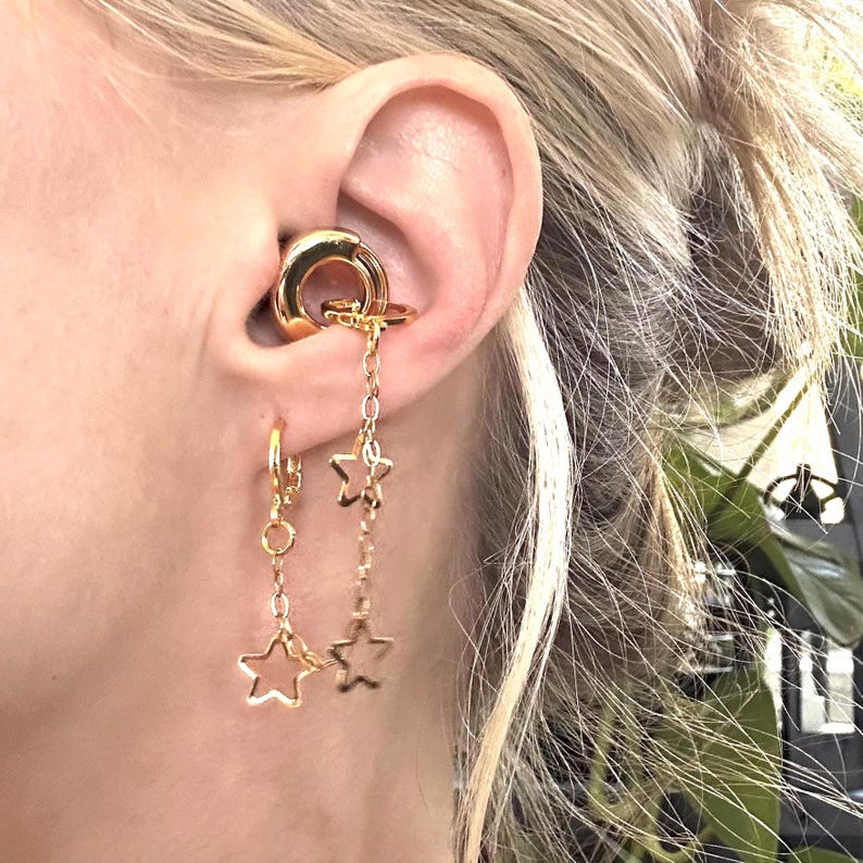 May include: A gold-toned ear cuff featuring a smooth circular band connected to a dangling chain with two open star charms, paired with a matching gold-toned hoop earring that has a single dangling open star charm attached to its base.