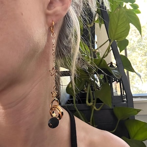 May include: A gold-toned dangle earring featuring a thin chain with two small star-shaped charms, ending in a gold-toned circular ring charm and a single matte black spherical bead. The earring is attached to the earlobe with a gold-toned hoop clasp.