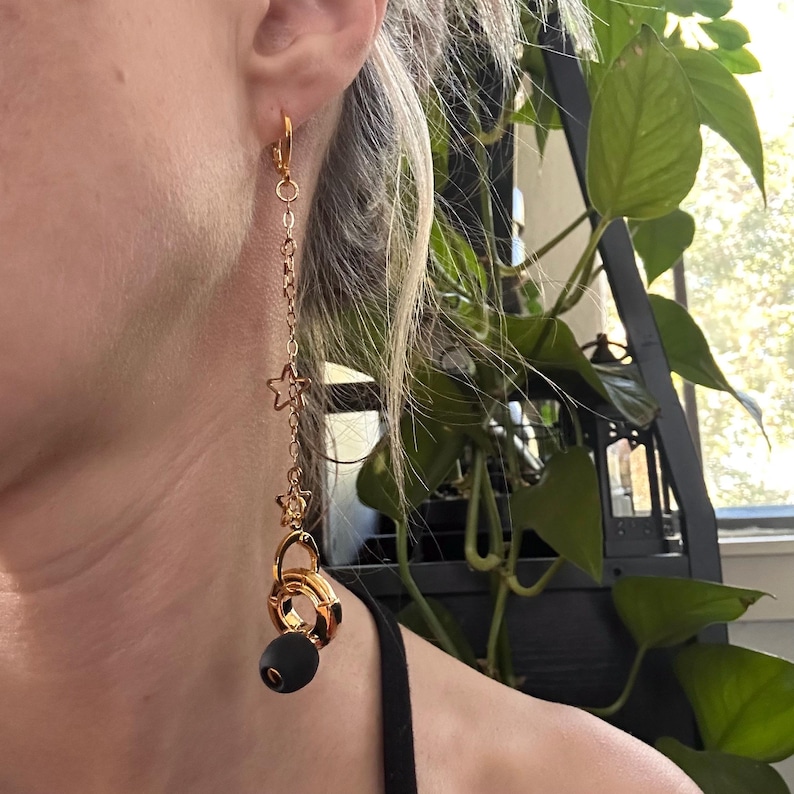 May include: A gold-toned dangle earring featuring a thin chain with two small star-shaped charms, ending in a gold-toned circular ring charm and a single matte black spherical bead. The earring is attached to the earlobe with a gold-toned hoop clasp.
