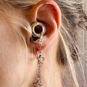 May include: A silver-toned ear gauge tunnel featuring a dangling chain attachment. The chain is adorned with two open-frame star charms. The jewelry is inserted into the earlobe, with the chain and star charms hanging vertically from the tunnel.
