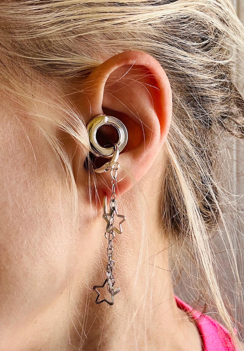 May include: A silver-toned ear gauge tunnel featuring a dangling chain attachment. The chain is adorned with two open-frame star charms. The jewelry is inserted into the earlobe, with the chain and star charms hanging vertically from the tunnel.