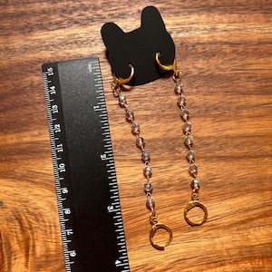 May include: A pair of gold-toned hoop earrings with dangling chains of iridescent beads attached to a black cat-shaped display card, positioned next to a black ruler showing a length of approximately 3.5 inches for the beaded chain section.