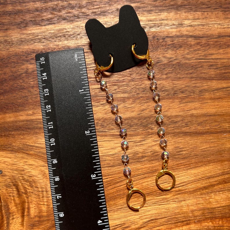 May include: A pair of gold-toned hoop earrings with dangling chains of iridescent beads attached to a black cat-shaped display card, positioned next to a black ruler showing a length of approximately 3.5 inches for the beaded chain section.