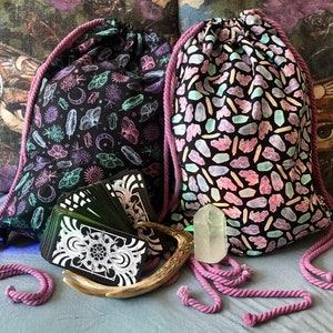 May include: Two drawstring bags with black fabric and different patterns. One bag has a pattern of crystals and the other has a pattern of moths, moons, and feathers. The bags are on a light blue surface with a deck of tarot cards and a white crystal.