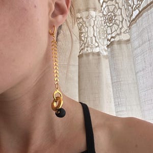 May include: A long gold-toned earring featuring a small hoop at the top, a vertical chain composed of repeating chevron-shaped links, and a dangling charm assembly consisting of two interlocking gold rings and a small, smooth matte black sphere.