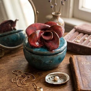 Peut inclure: Une boîte à bijoux en céramique turquoise avec un couvercle en forme de fleur rouge est posée sur une surface en bois. Un collier avec un pendentif en forme de cœur doré, une petite coquille avec une perle et un livre vintage sont également visibles.