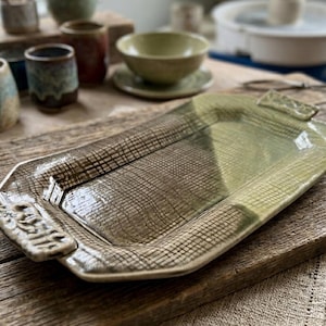 Peut inclure: Un plateau de service en céramique fait à la main, avec une surface texturée et une forme rectangulaire. Le plateau présente un mélange de tons vert olive et marron, avec des poignées décoratives à chaque extrémité. D'autres pièces en céramique sont en arrière-plan.