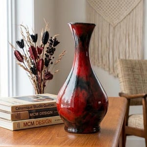 May include: A tall, bulbous ceramic vase with a glossy finish featuring a marbled pattern of deep red and black. The vase has a narrow neck that flares slightly at the rim and a rounded base. It is positioned next to a stack of three books.