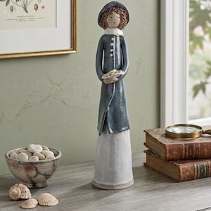 May include: A ceramic figurine of a person in a long blue coat and hat, holding a small dish. The figurine has a white skirt and curly brown hair. The figurine is on a wooden surface with a bowl of stones and seashells.
