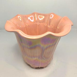 Peut inclure: Un vase en céramique pêche irisé avec un bord festonné. Le vase a une forme de tulipe avec des rainures verticales et un éclat nacré, reflétant des teintes de rose, de jaune et de violet. La base a trois petits pieds.