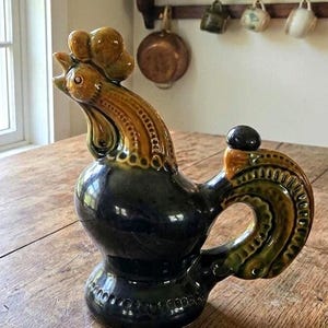 May include: A ceramic rooster-shaped pitcher with a black base and a brown and green neck and head. The pitcher has a handle and a small black stopper on top. It is sitting on a wooden table.