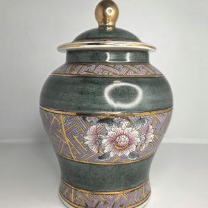 Peut inclure: Un pot décoratif en céramique avec un couvercle, de couleur vert foncé. Le pot est orné d'une bordure dorée et d'un motif floral rose et blanc. Le couvercle est surmonté d'un bouton doré.