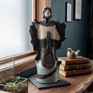 May include: A ceramic sculpture of a figure with a mandolin. The sculpture is primarily black, cream, and blue-green. It sits on a dark square base. The background includes books, a decorative bird, and framed artwork.
