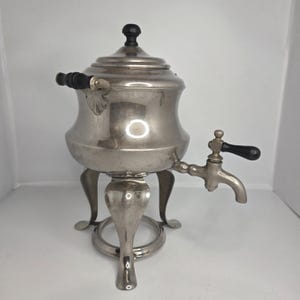 May include: A vintage silver-toned coffee urn with a black handle and spout. The urn has a rounded body, a lid with a black knob, and sits on three decorative legs. The spout has a black handle.
