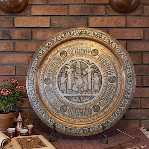 Puede incluir: Una gran bandeja de cobre ornamentada con intrincados diseños grabados, incluyendo figuras y caligrafía. La bandeja se exhibe sobre un soporte negro contra una pared de ladrillo, con apliques de cobre encima.